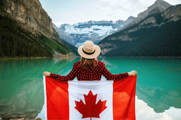 Woman Wearing Red and Black Checkered Dress Shirt and Beige Fedora Hat Holding Canada Flag Looking at Lake
