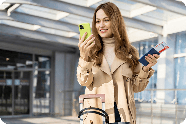 A traveler in a beige trench coat holds a green phone and a passport while standing next to a suitcase in a modern airport setting