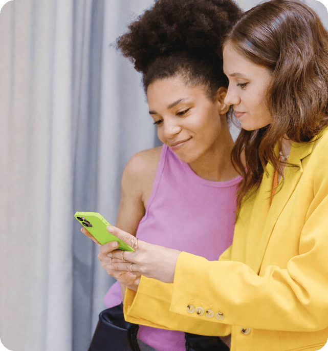 Two women closely examine a smartphone, one wearing a vibrant yellow jacket and the other in a pink top, with a soft background